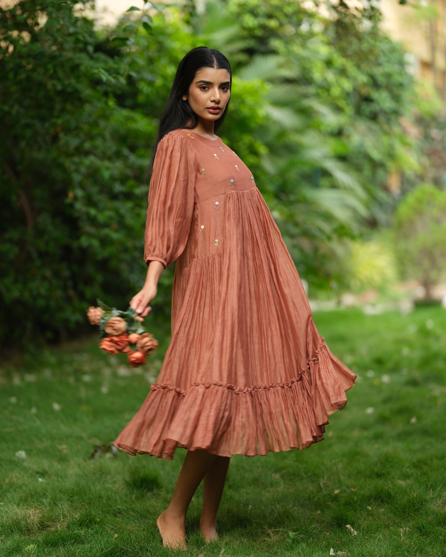 Woman wearing a rust-colored Mul Chanderi smocked midi dress with floral embroidery, standing barefoot in a green garden.