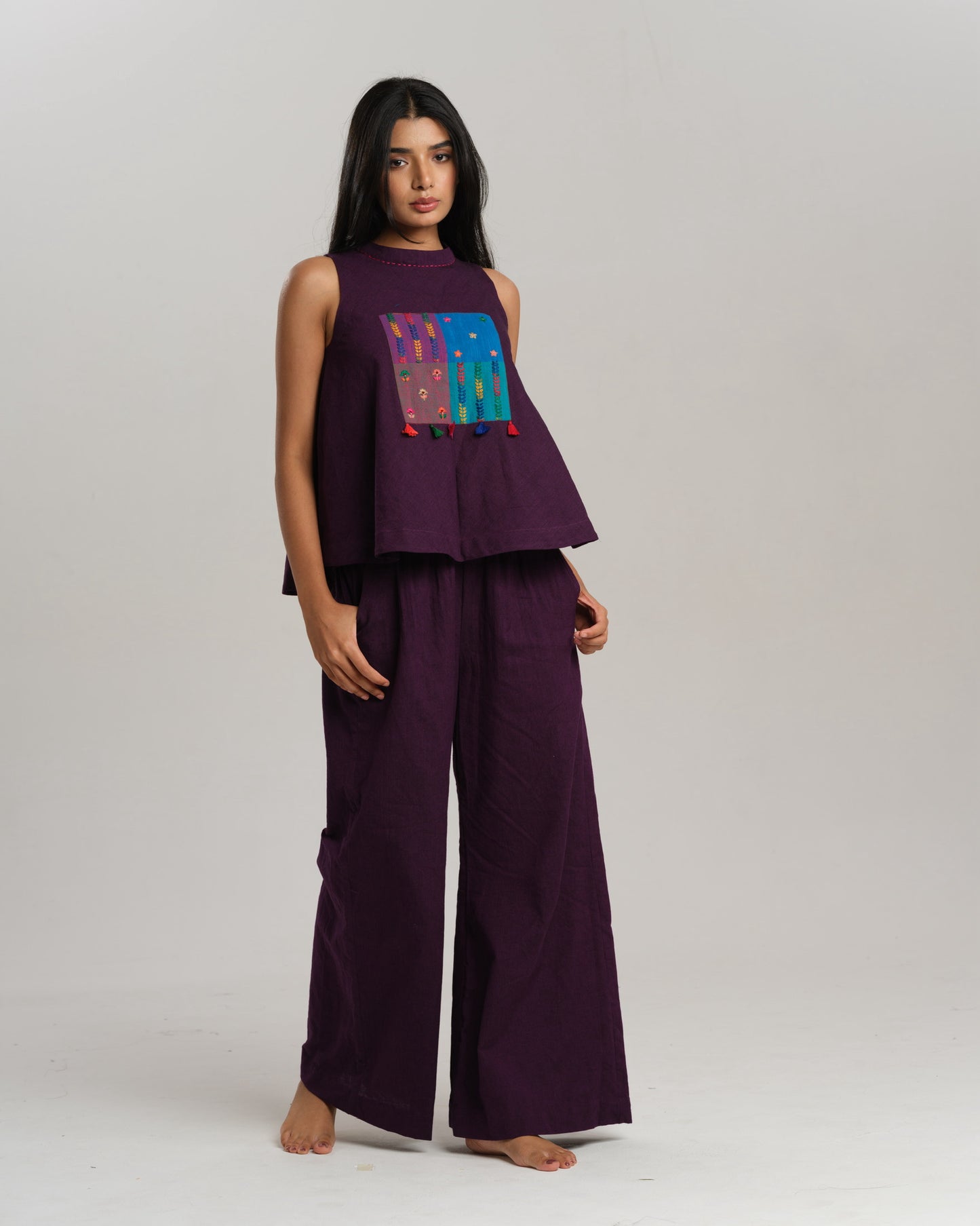 Model wearing a deep plum ikat co-ord set with a sleeveless flared top featuring colourful handwoven ikat patchwork and wide-leg pants, standing barefoot against a neutral background.