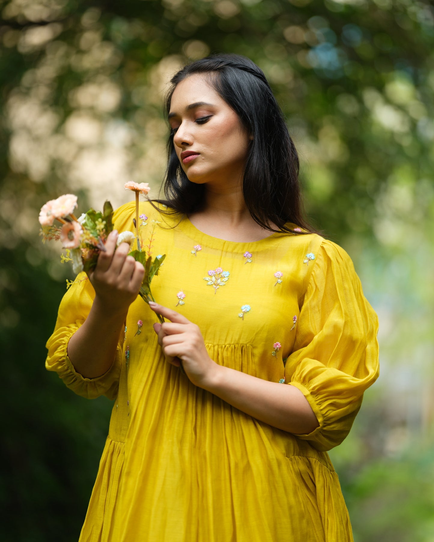 Model wearing a flowy yellow mul Chanderi embroidered dress, holding flowers and standing on green grass.