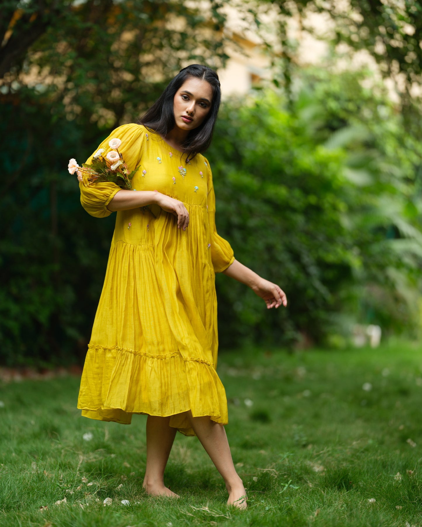 Model wearing a flowy yellow mul Chanderi embroidered dress, holding flowers and standing on green grass.