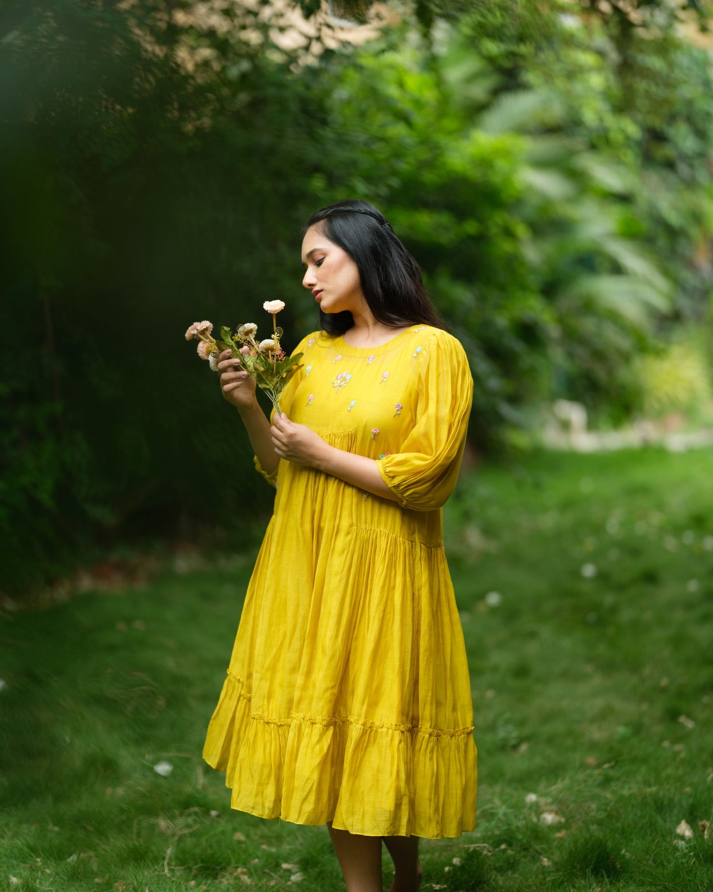 Model wearing a flowy yellow mul Chanderi embroidered dress, holding flowers and standing on green grass.