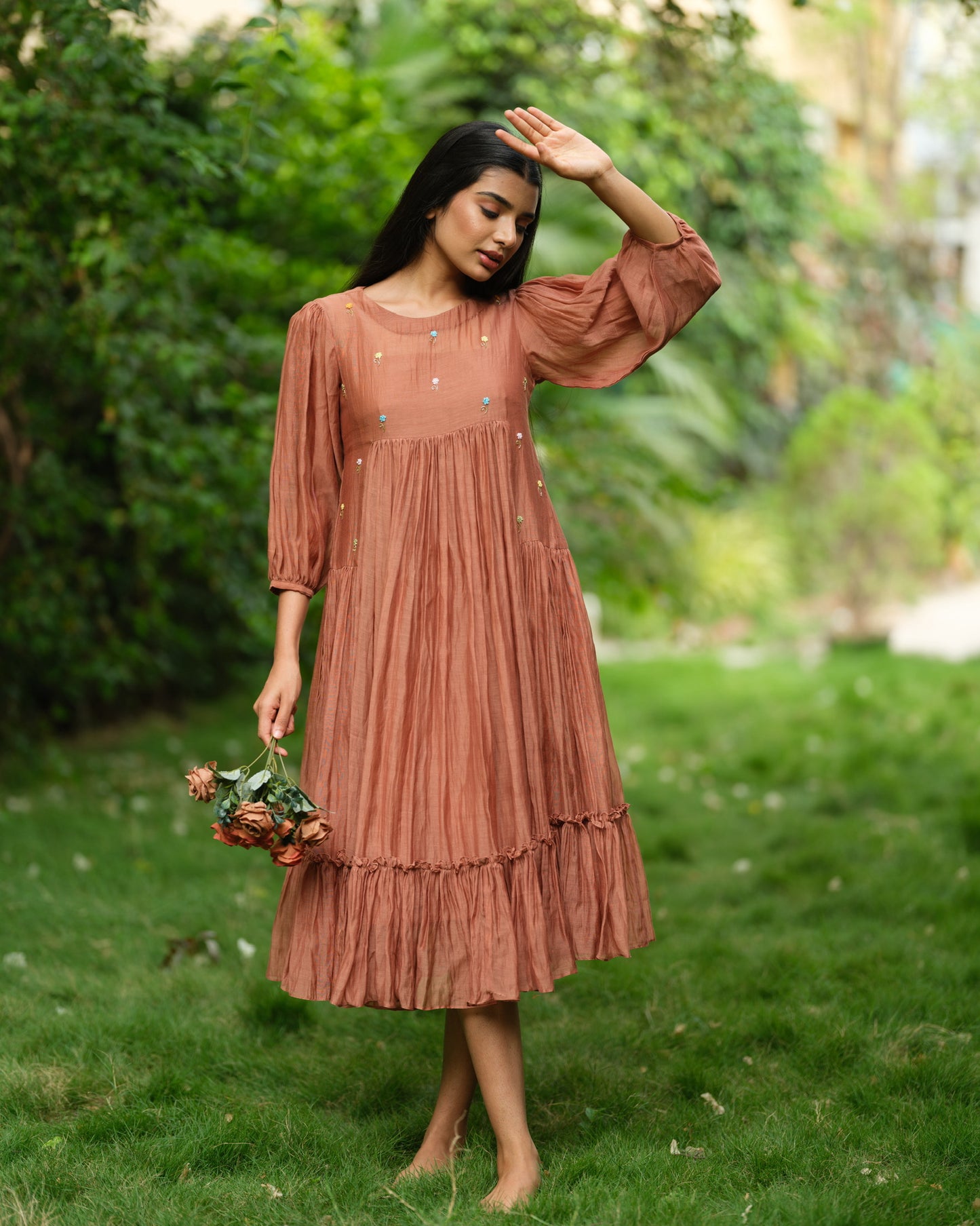 Woman wearing a rust-colored Mul Chanderi smocked midi dress with floral embroidery, standing barefoot in a green garden.