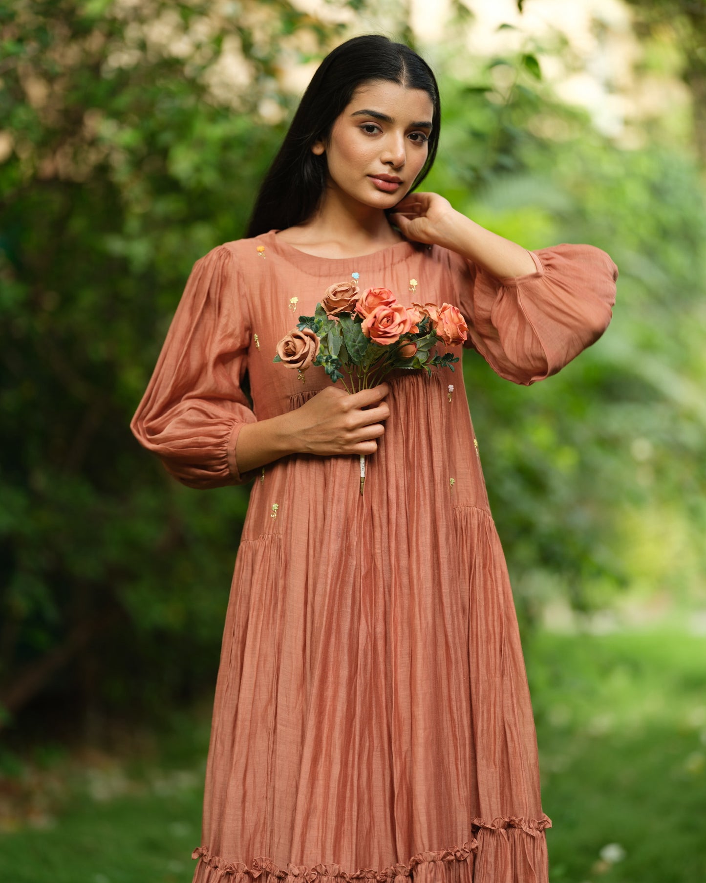 Woman wearing a rust-colored Mul Chanderi smocked midi dress with floral embroidery, standing barefoot in a green garden.