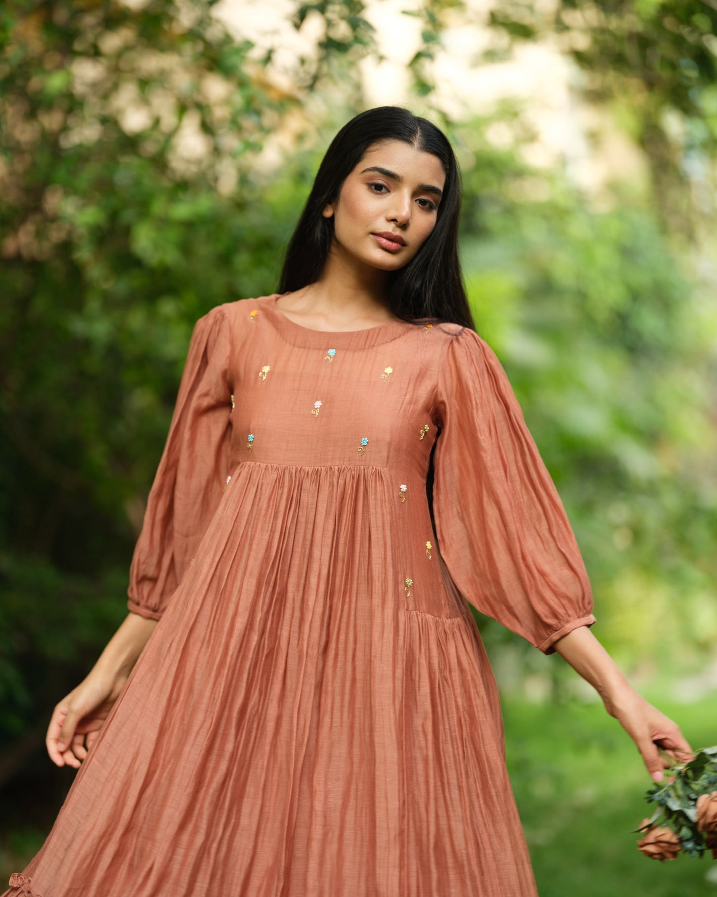Woman wearing a rust-colored Mul Chanderi smocked midi dress with floral embroidery, standing barefoot in a green garden.