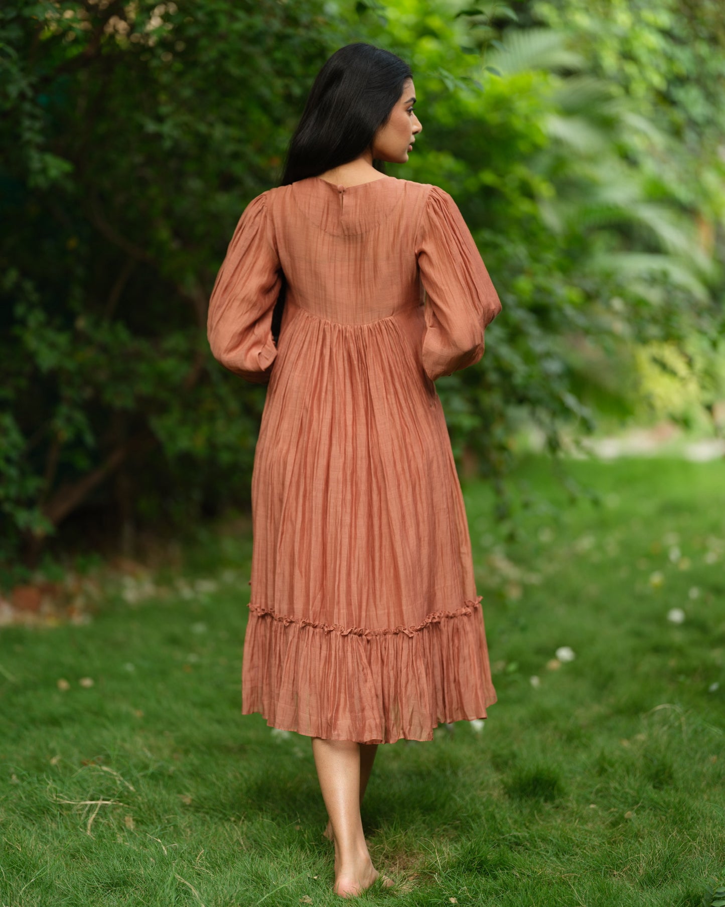Woman wearing a rust-colored Mul Chanderi smocked midi dress with floral embroidery, standing barefoot in a green garden.