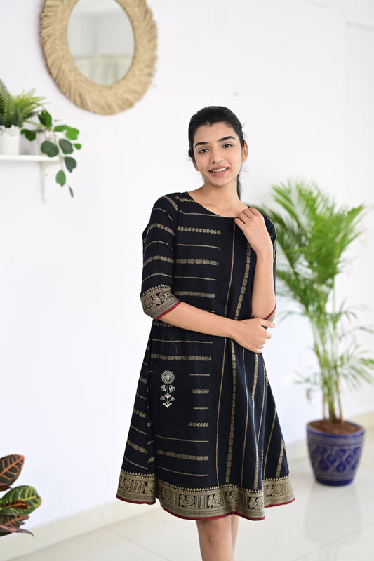 Model wearing a black fusion Narayanpet handmade dress with intricate handloom patterns, featuring a body-con silhouette and elegant motifs, standing in a minimalist indoor setting with plants in the background