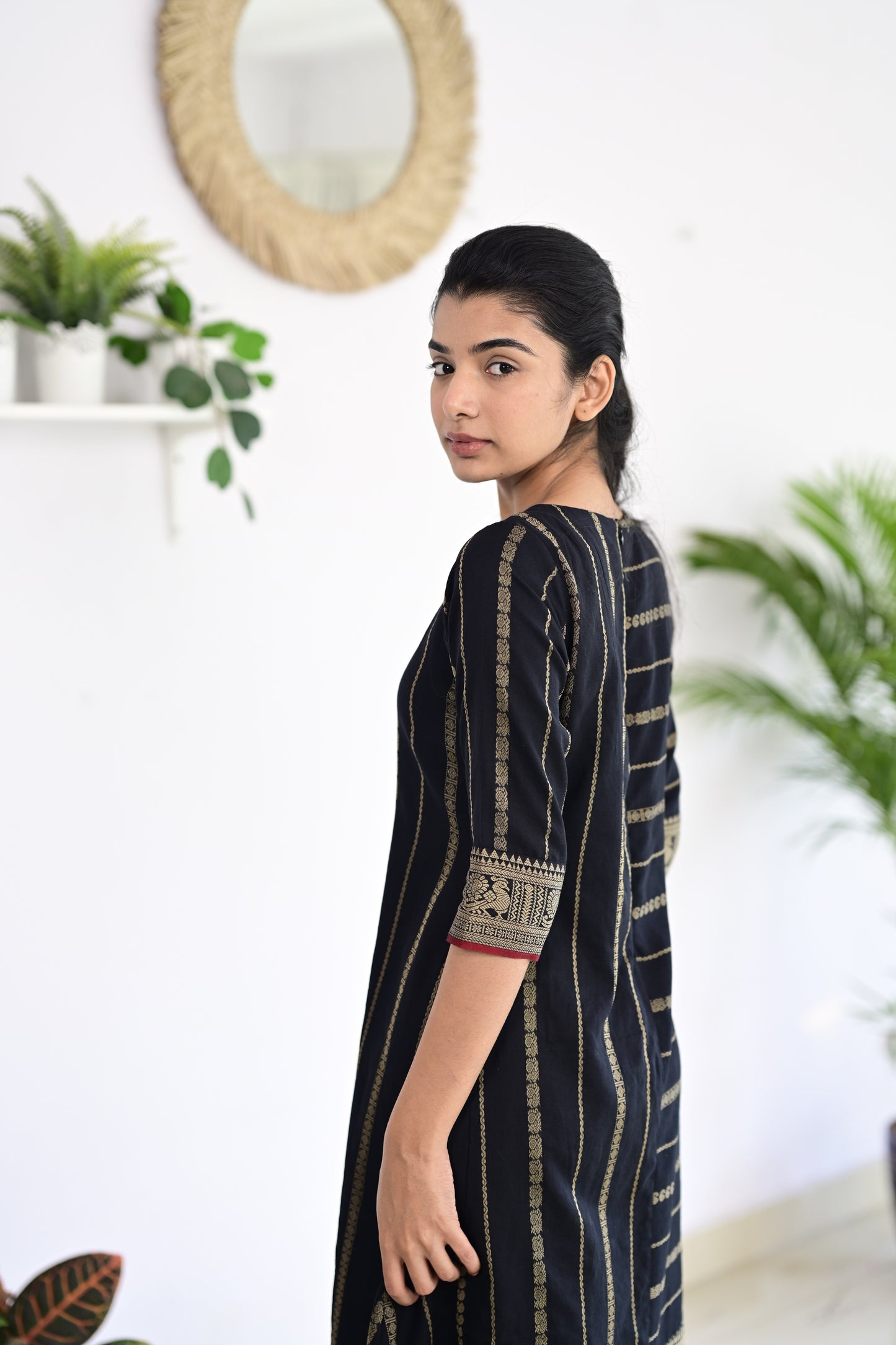 Model wearing a black fusion Narayanpet handmade dress with intricate handloom patterns, featuring a body-con silhouette and elegant motifs, standing in a minimalist indoor setting with plants in the background