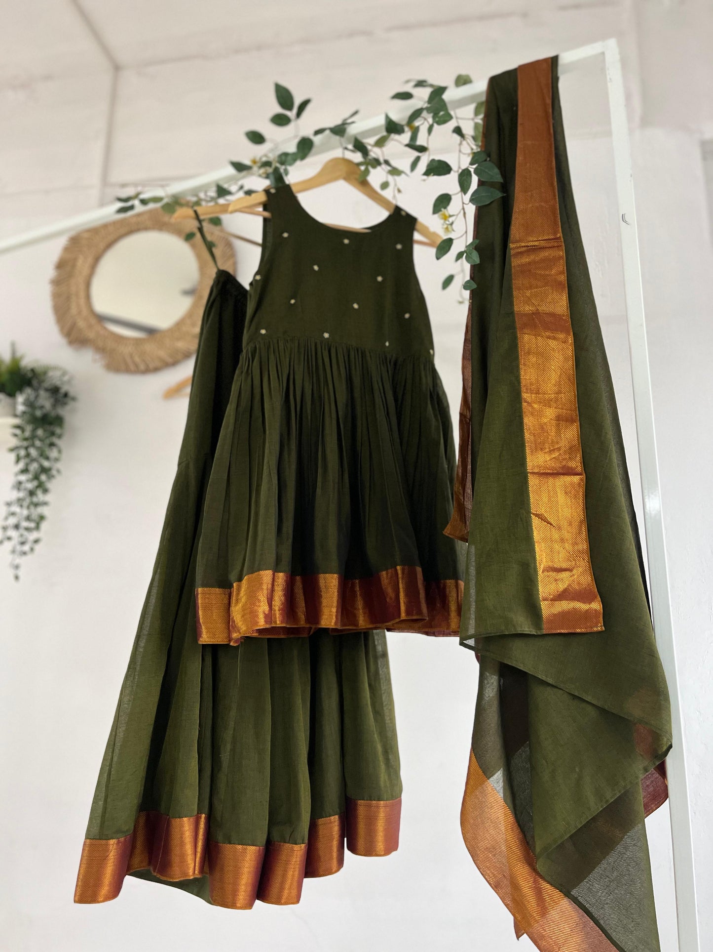Olive green Narayanpet sharara set with golden zari borders, flared kurta, sharara pants, and dupatta displayed on hangers against a white backdrop with greenery décor.