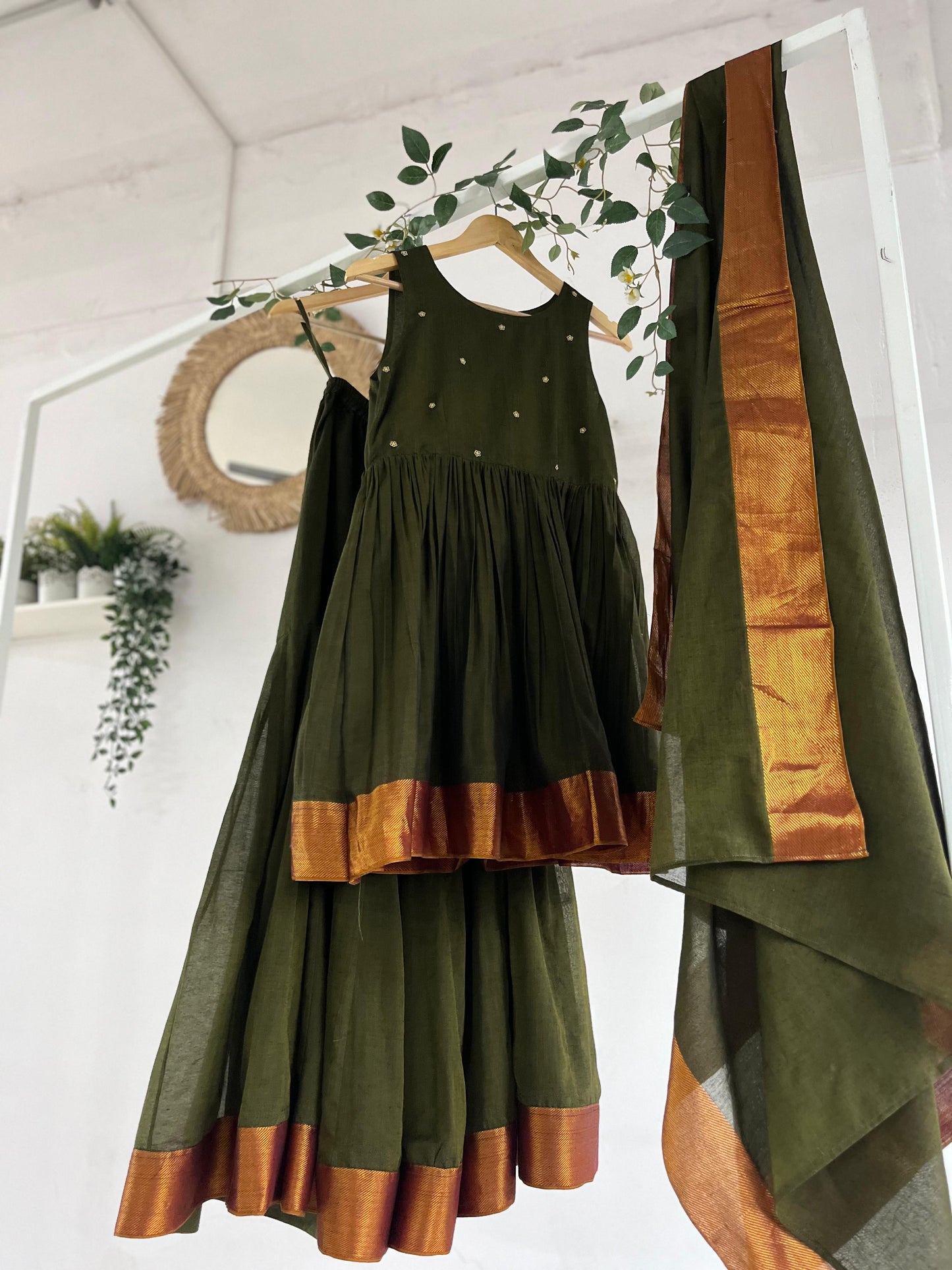 Olive green Narayanpet sharara set with golden zari borders, flared kurta, sharara pants, and dupatta displayed on hangers against a white backdrop with greenery décor.