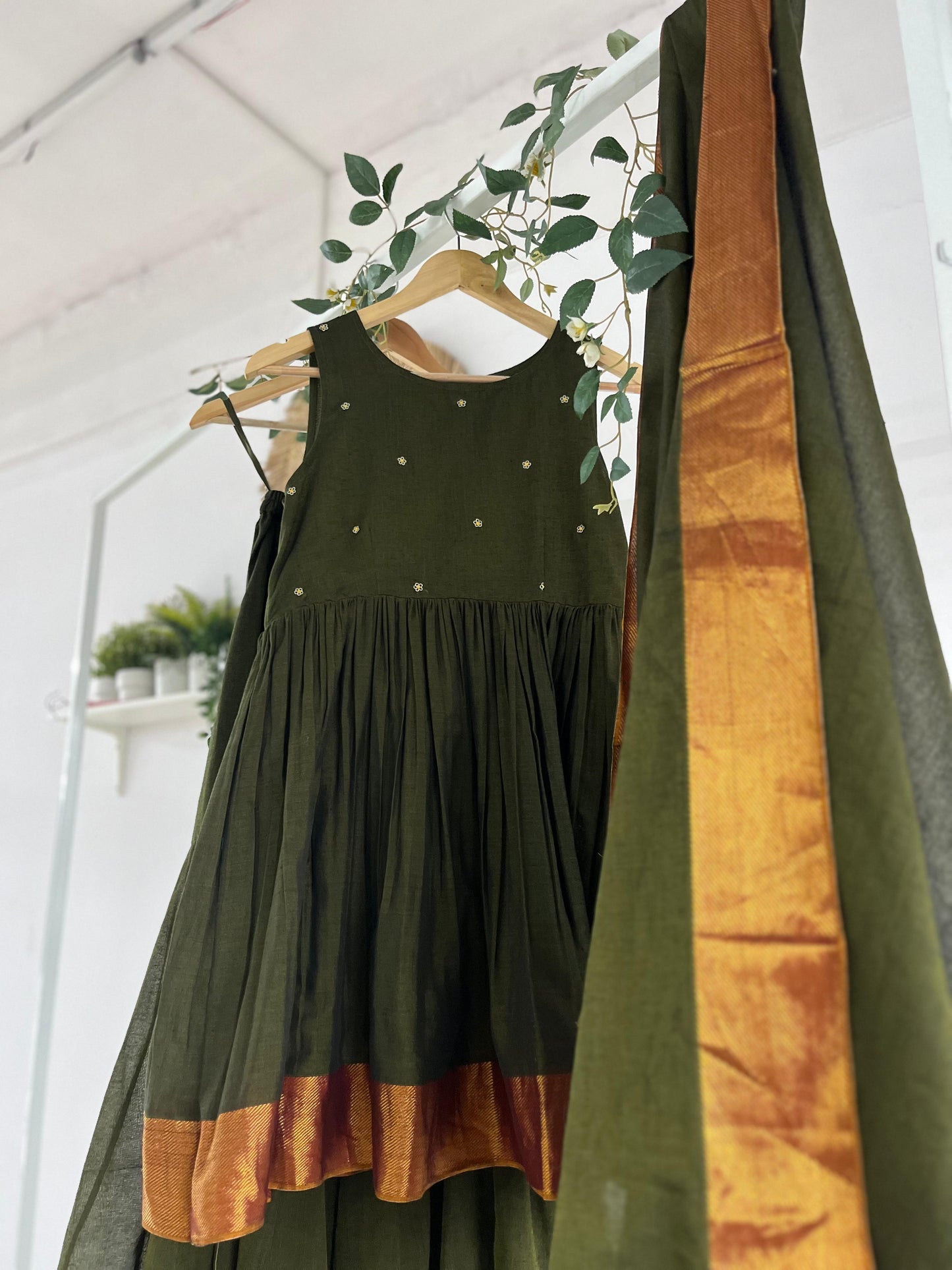 Olive green Narayanpet sharara set with golden zari borders, flared kurta, sharara pants, and dupatta displayed on hangers against a white backdrop with greenery décor.