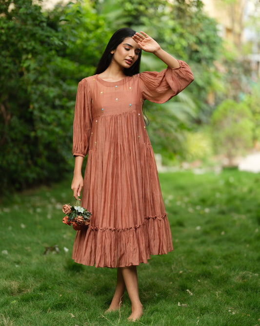 Woman wearing a rust-colored Mul Chanderi smocked midi dress with floral embroidery, standing barefoot in a green garden.