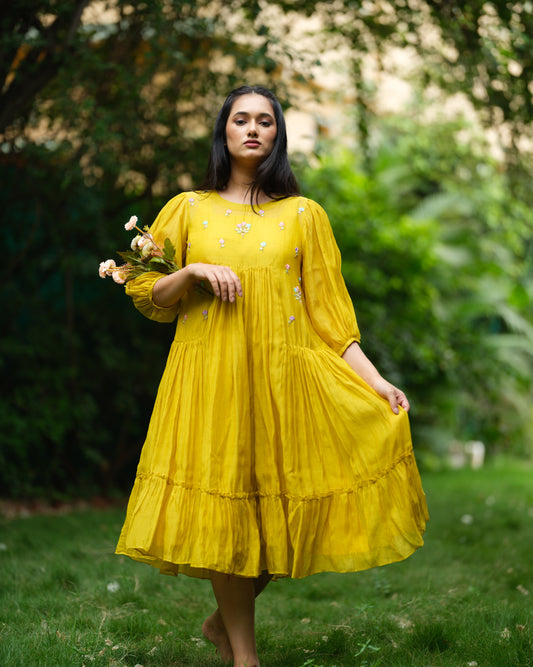 Model wearing a flowy yellow mul Chanderi embroidered dress, holding flowers and standing on green grass.