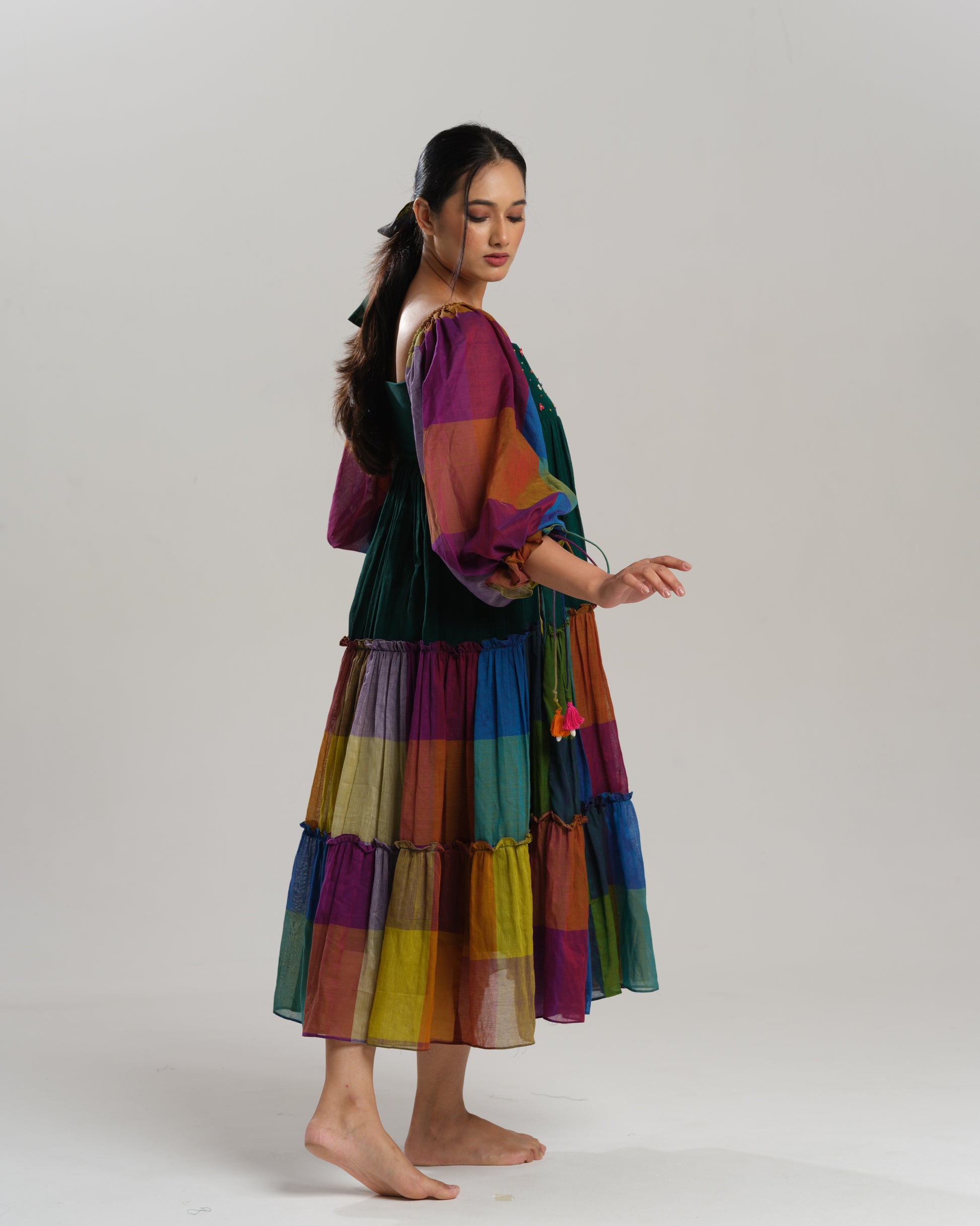 Model wearing a multicoloured handwoven Narayanpet checks cotton tiered dress with puff sleeves and mirror work bodice, styled against a minimalist background.