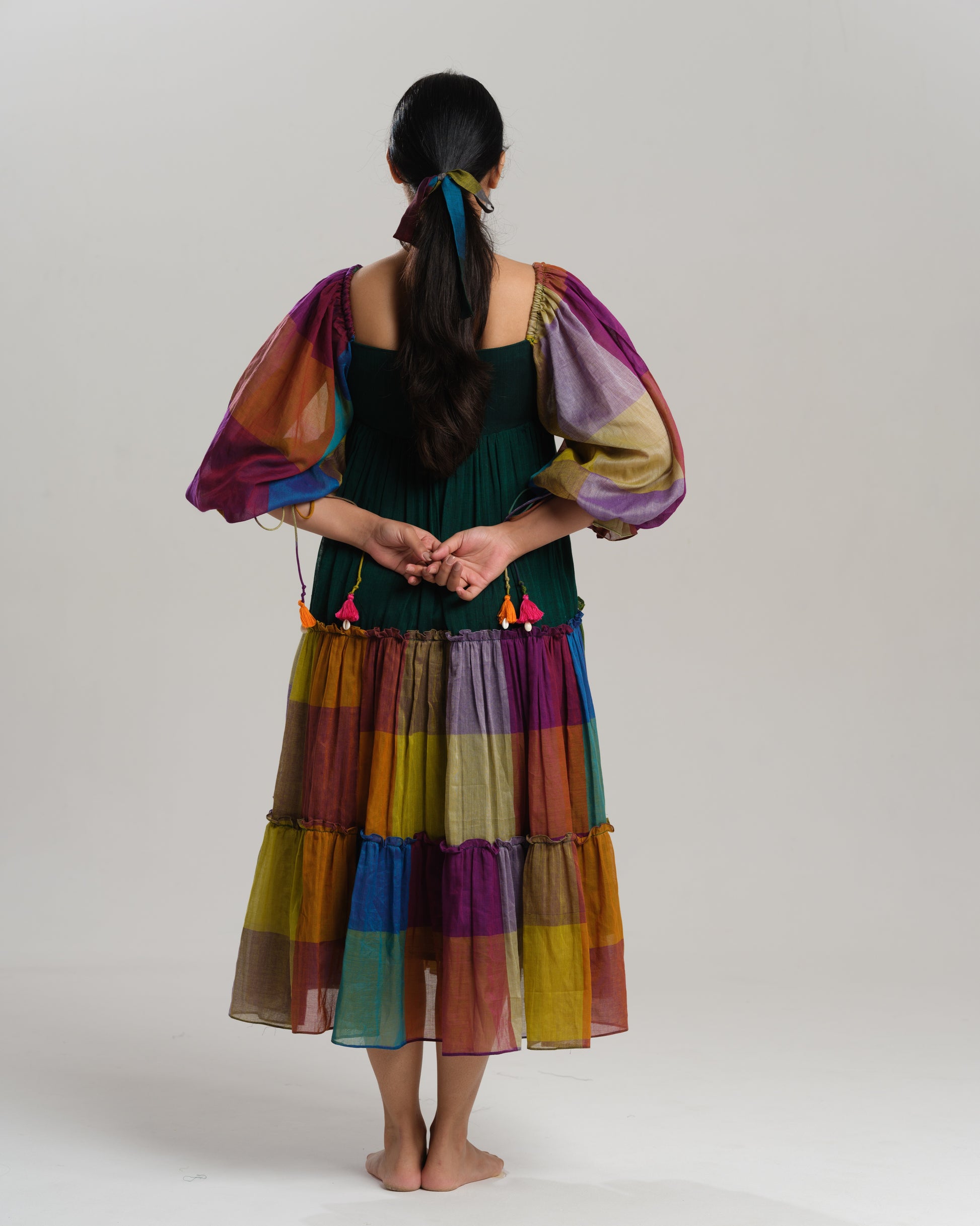Model wearing a multicoloured handwoven Narayanpet checks cotton tiered dress with puff sleeves and mirror work bodice, styled against a minimalist background.