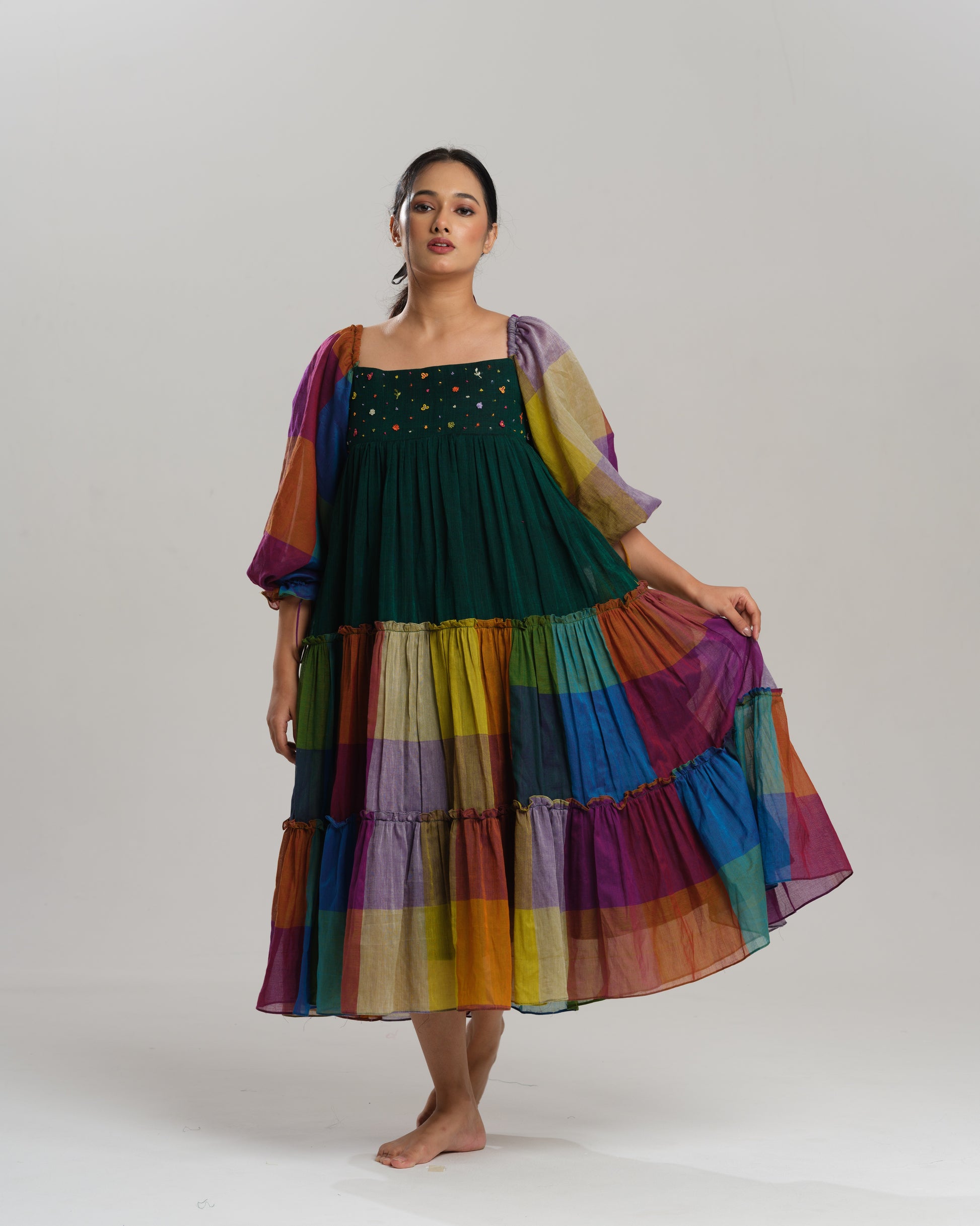 Model wearing a multicoloured handwoven Narayanpet checks cotton tiered dress with puff sleeves and mirror work bodice, styled against a minimalist background.