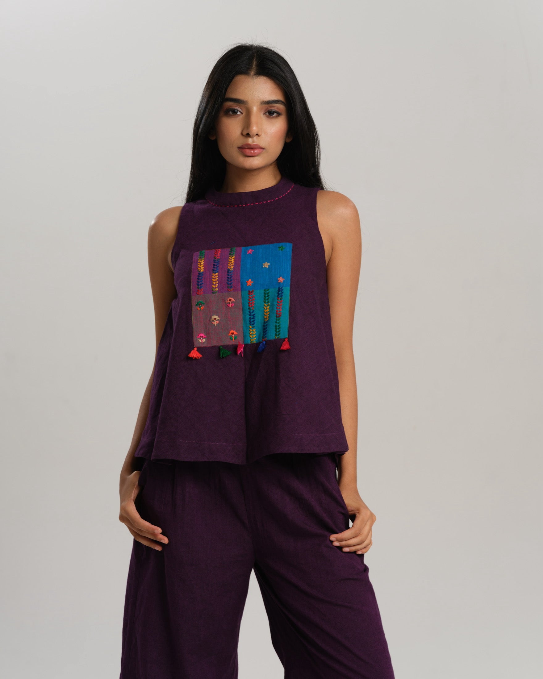 Model wearing a deep plum ikat co-ord set with a sleeveless flared top featuring colourful handwoven ikat patchwork and wide-leg pants, standing barefoot against a neutral background.