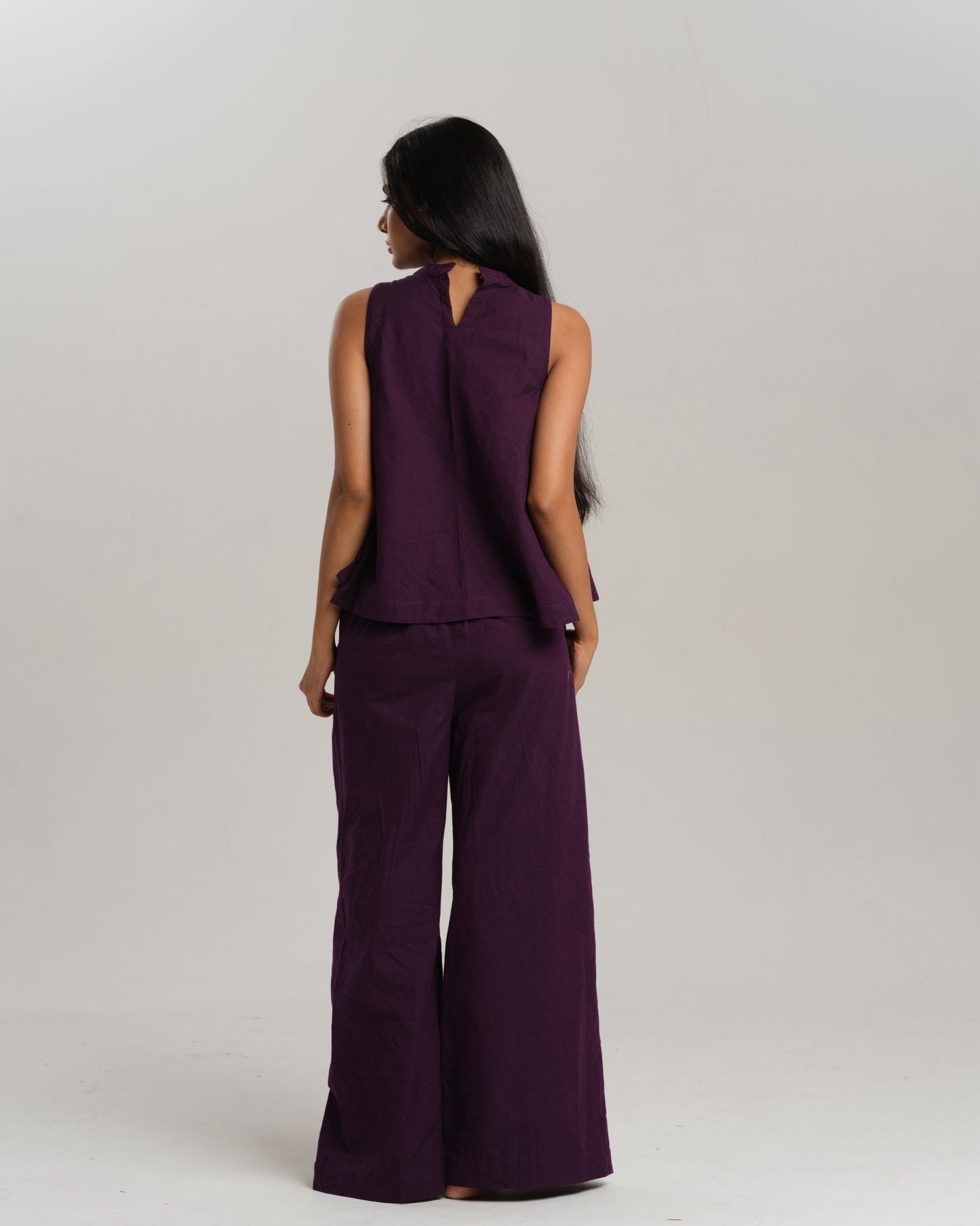 Model wearing a deep plum ikat co-ord set with a sleeveless flared top featuring colourful handwoven ikat patchwork and wide-leg pants, standing barefoot against a neutral background.