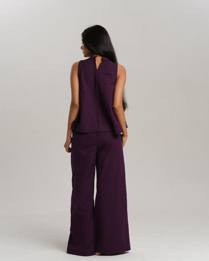 Model wearing a deep plum ikat co-ord set with a sleeveless flared top featuring colourful handwoven ikat patchwork and wide-leg pants, standing barefoot against a neutral background.