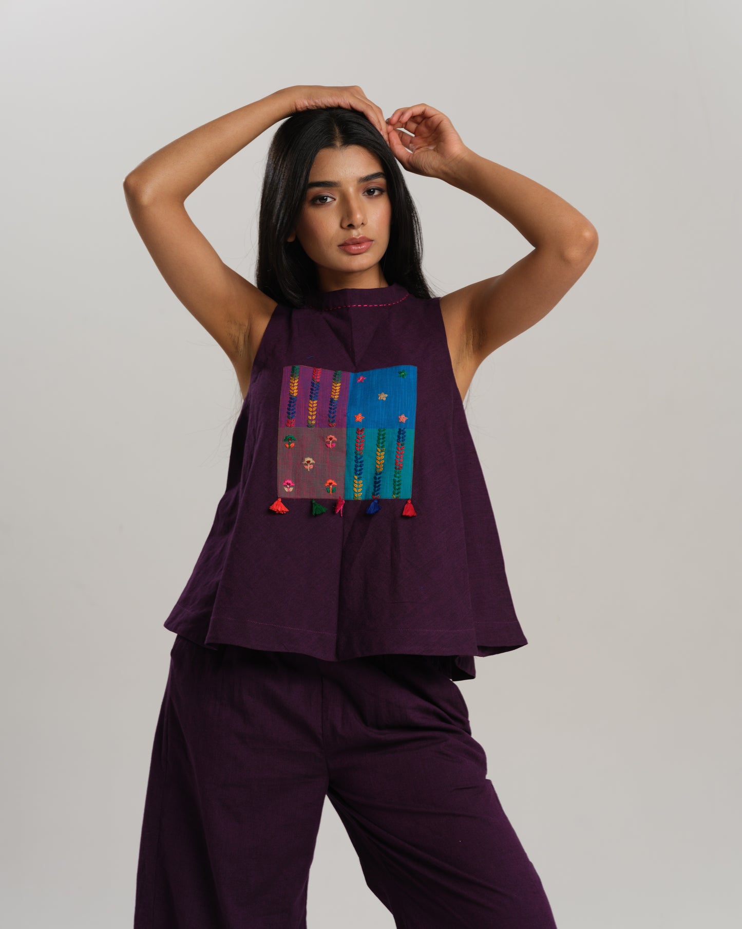 Model wearing a deep plum ikat co-ord set with a sleeveless flared top featuring colourful handwoven ikat patchwork and wide-leg pants, standing barefoot against a neutral background.