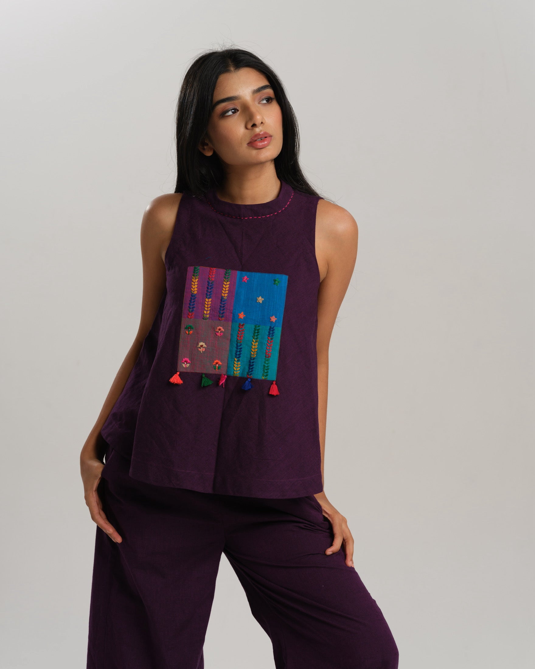 Model wearing a deep plum ikat co-ord set with a sleeveless flared top featuring colourful handwoven ikat patchwork and wide-leg pants, standing barefoot against a neutral background.