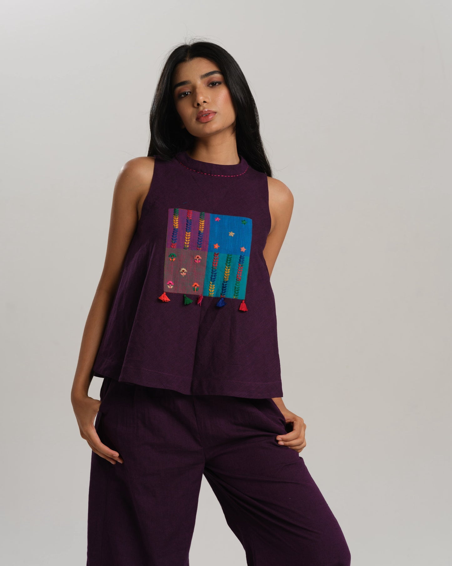 Model wearing a deep plum ikat co-ord set with a sleeveless flared top featuring colourful handwoven ikat patchwork and wide-leg pants, standing barefoot against a neutral background.