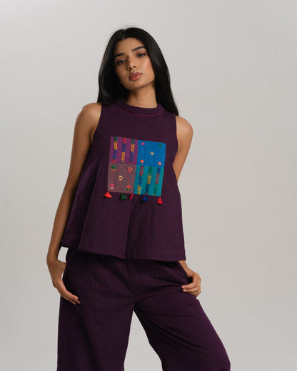 Model wearing a deep plum ikat co-ord set with a sleeveless flared top featuring colourful handwoven ikat patchwork and wide-leg pants, standing barefoot against a neutral background.