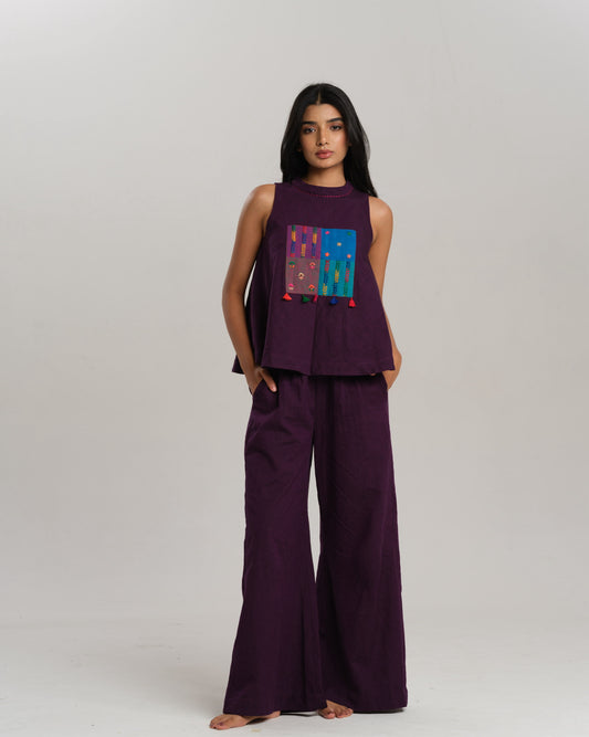 Model wearing a deep plum ikat co-ord set with a sleeveless flared top featuring colourful handwoven ikat patchwork and wide-leg pants, standing barefoot against a neutral background.