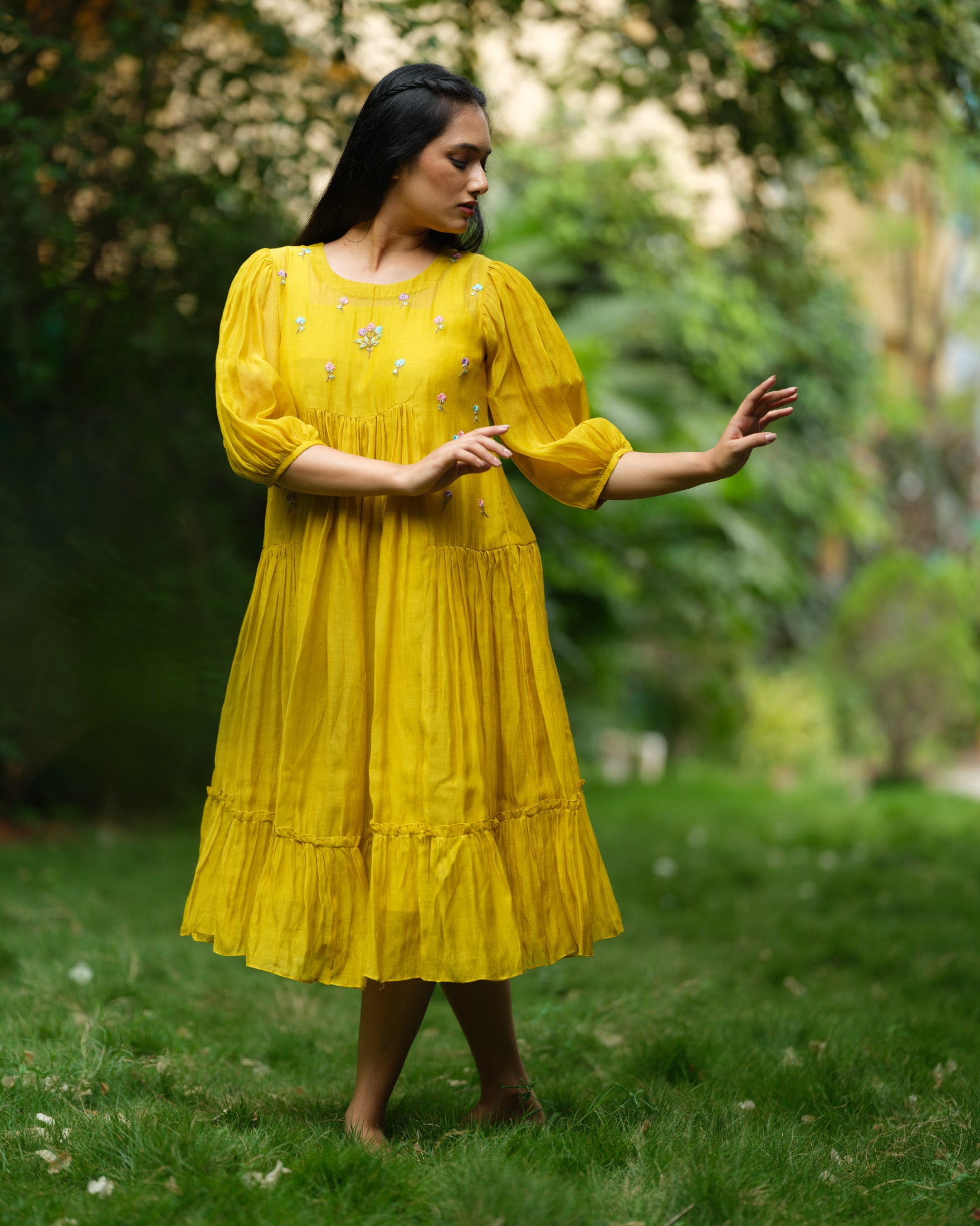 Model wearing a flowy yellow mul Chanderi embroidered dress, holding flowers and standing on green grass.