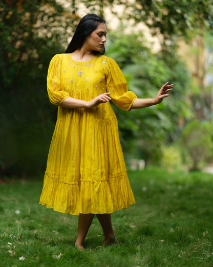 Model wearing a flowy yellow mul Chanderi embroidered dress, holding flowers and standing on green grass.