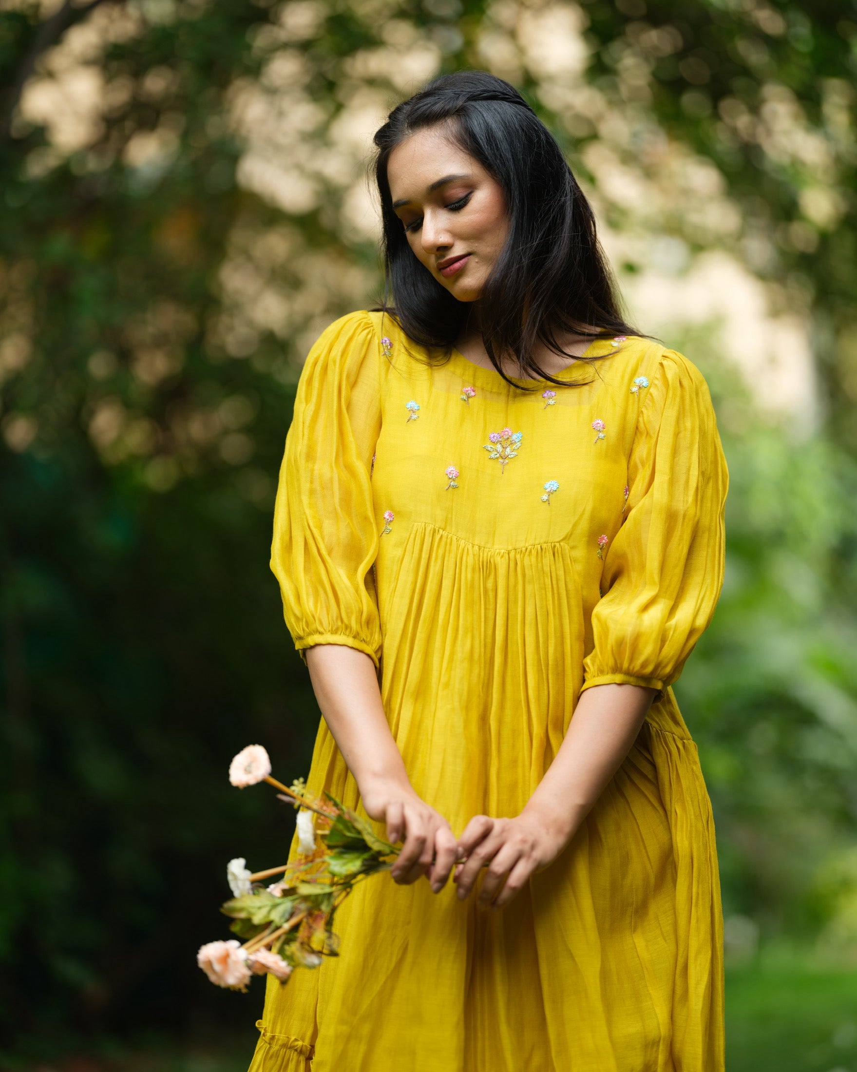 Model wearing a flowy yellow mul Chanderi embroidered dress, holding flowers and standing on green grass.
