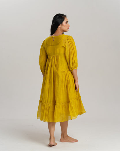 Model wearing a flowy yellow mul Chanderi embroidered dress, holding flowers and standing on green grass.
