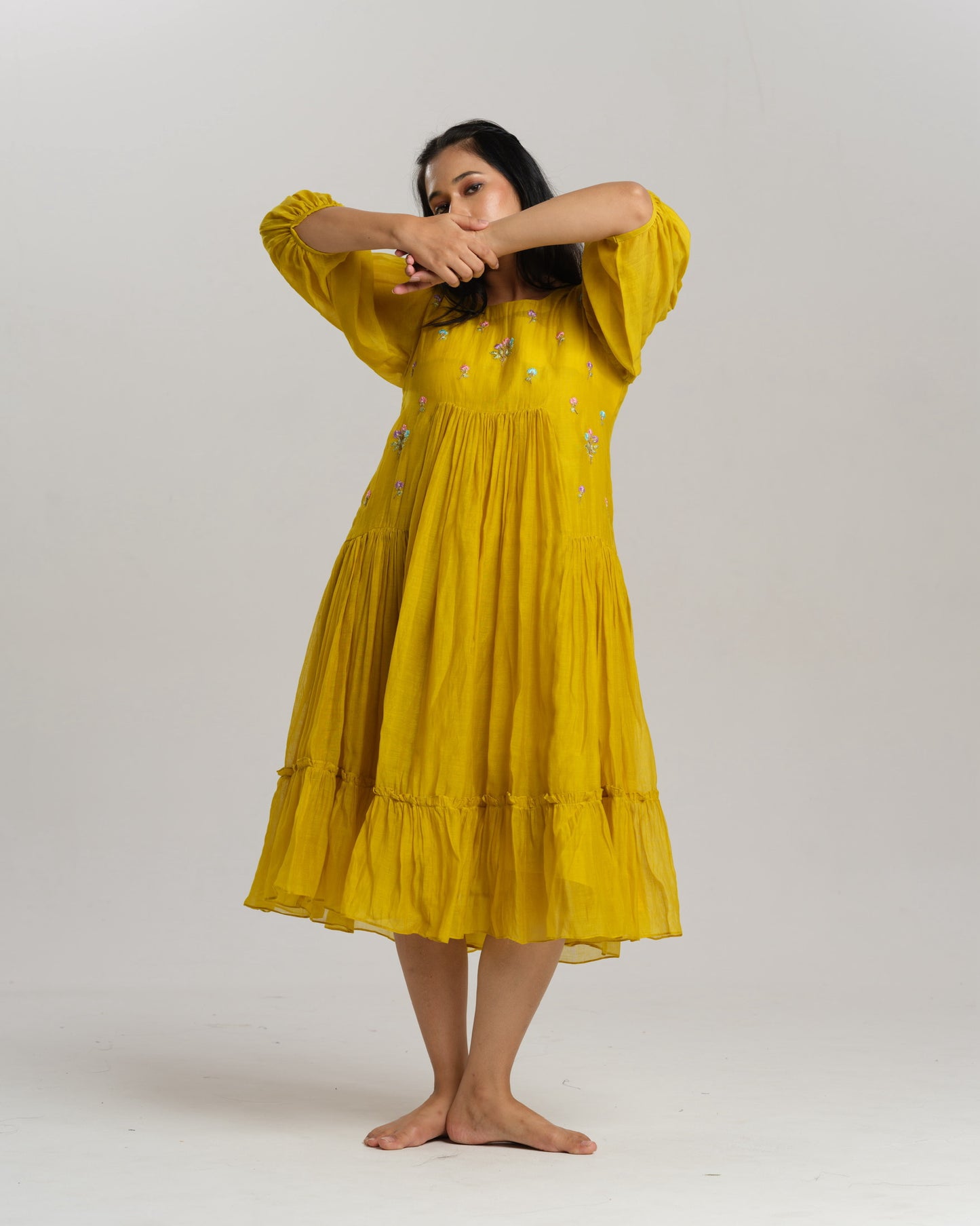 Model wearing a flowy yellow mul Chanderi embroidered dress, holding flowers and standing on green grass.