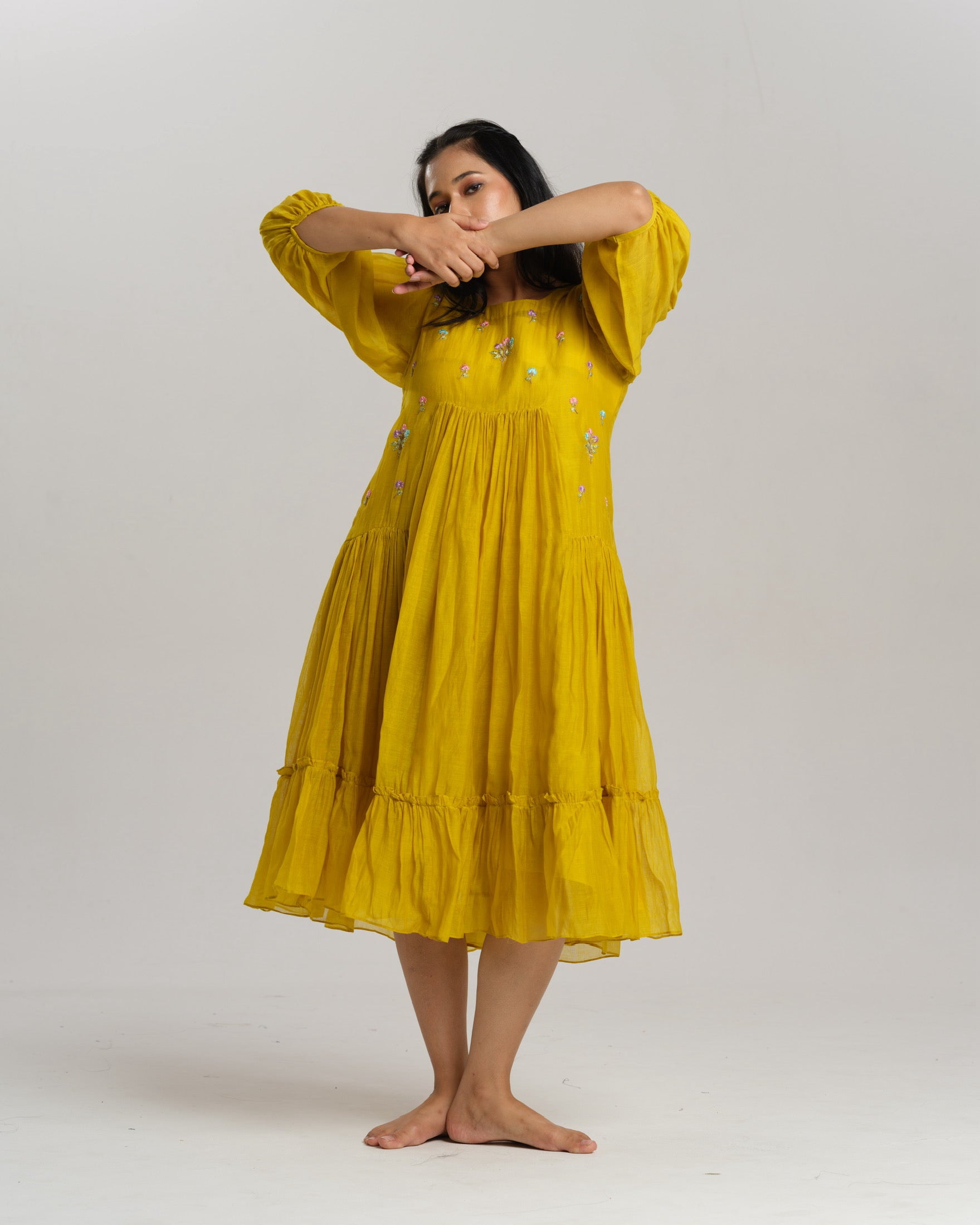 Model wearing a flowy yellow mul Chanderi embroidered dress, holding flowers and standing on green grass.