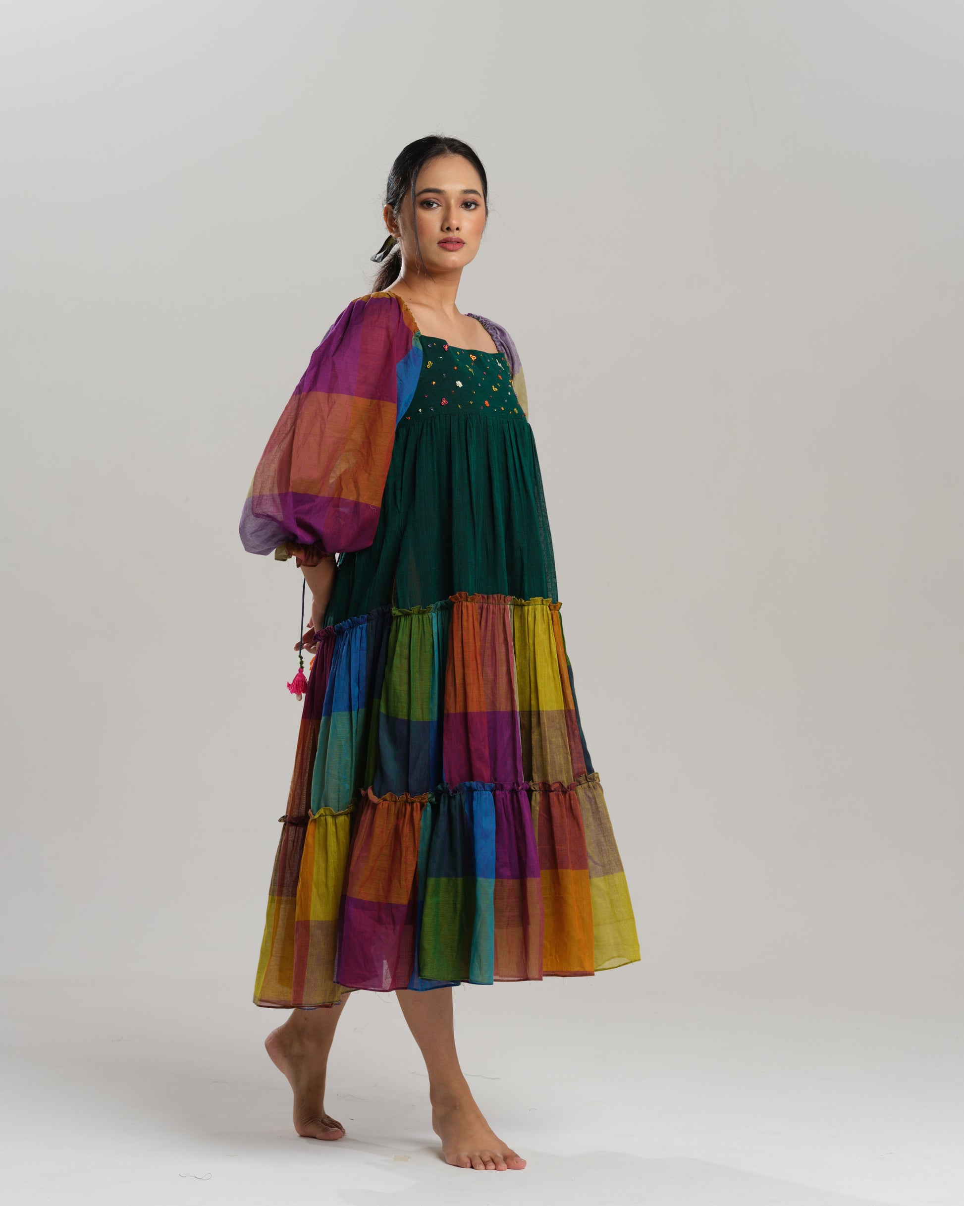 Model wearing a multicoloured handwoven Narayanpet checks cotton tiered dress with puff sleeves and mirror work bodice, styled against a minimalist background.