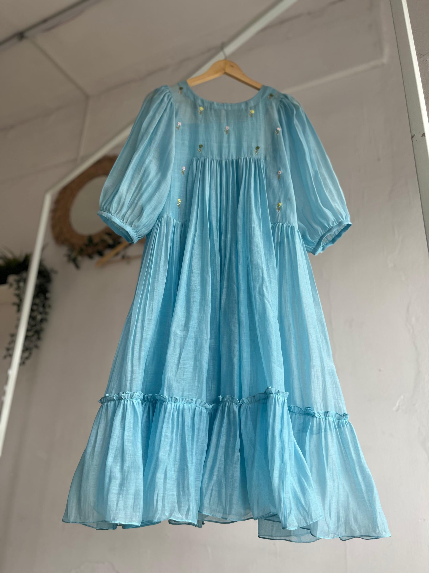 Powder blue cotton mul dress with puff sleeves, floral hand embroidery on the yoke, and a two-tiered flared hem hanging on a wooden hanger against a white background.