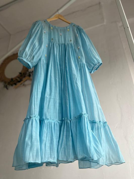Powder blue cotton mul dress with puff sleeves, floral hand embroidery on the yoke, and a two-tiered flared hem hanging on a wooden hanger against a white background.