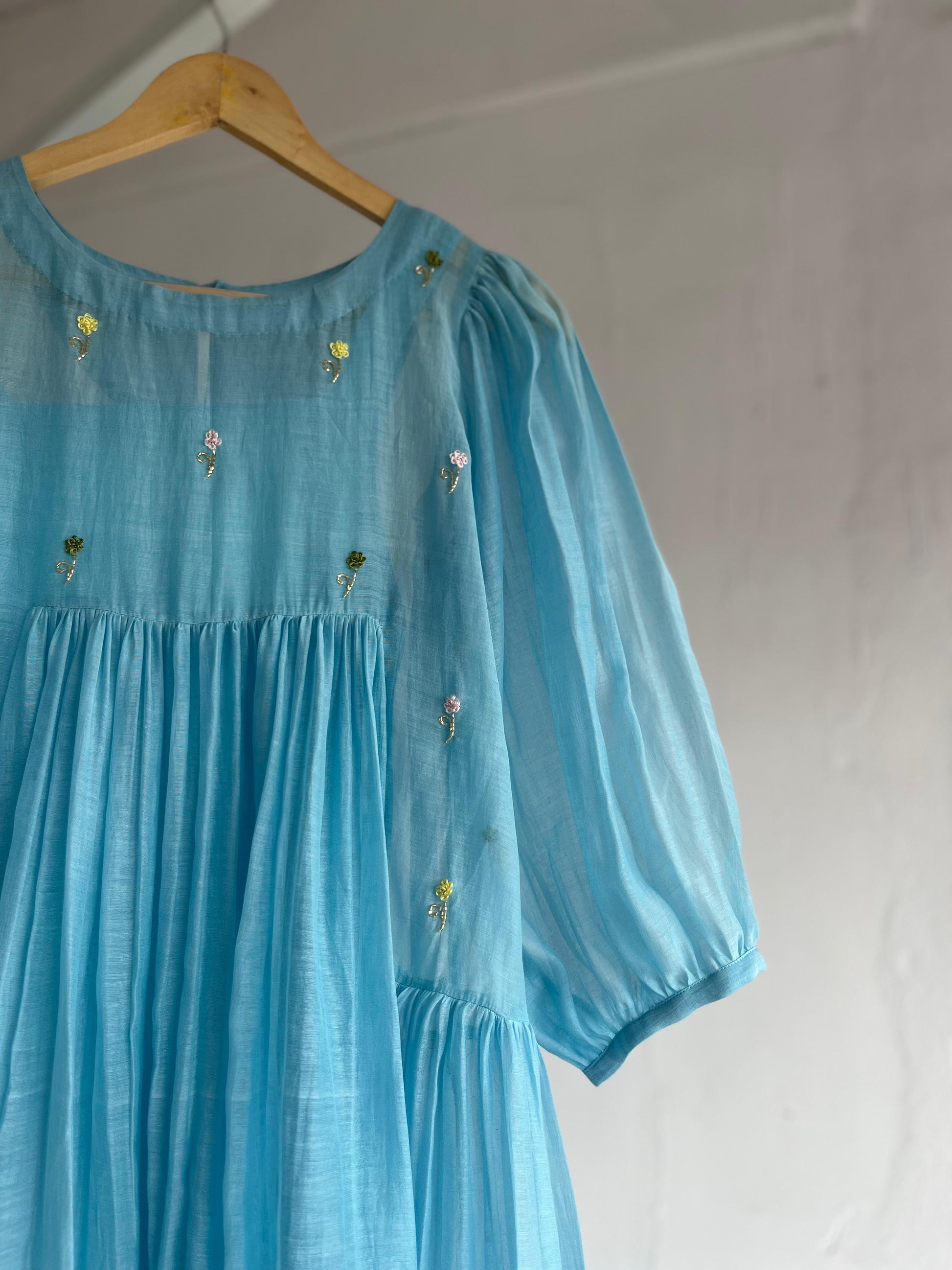 Powder blue cotton mul dress with puff sleeves, floral hand embroidery on the yoke, and a two-tiered flared hem hanging on a wooden hanger against a white background.