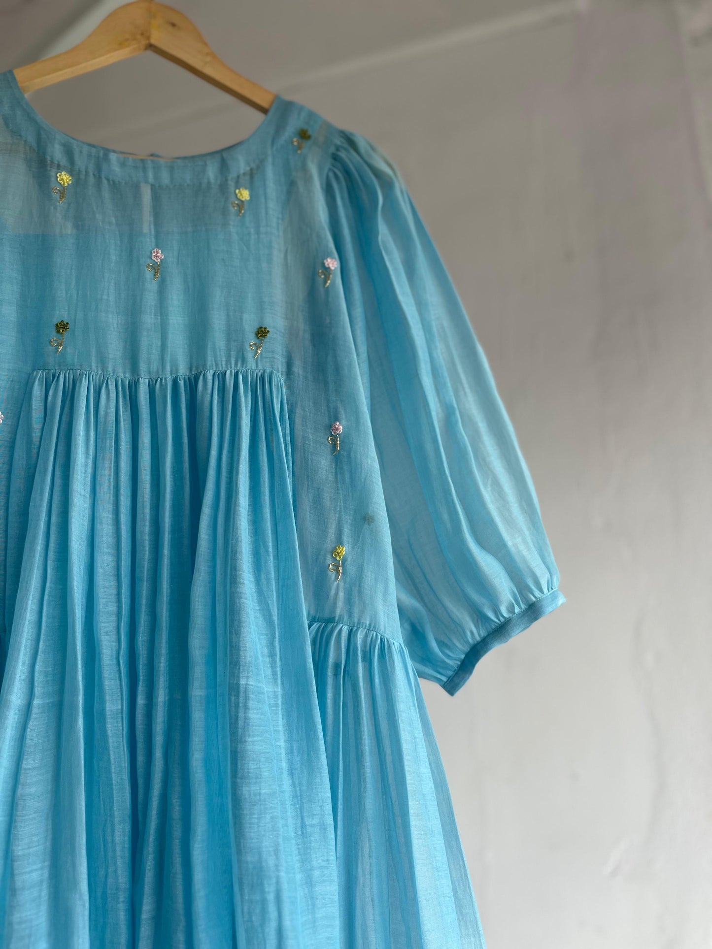 Powder blue cotton mul dress with puff sleeves, floral hand embroidery on the yoke, and a two-tiered flared hem hanging on a wooden hanger against a white background.