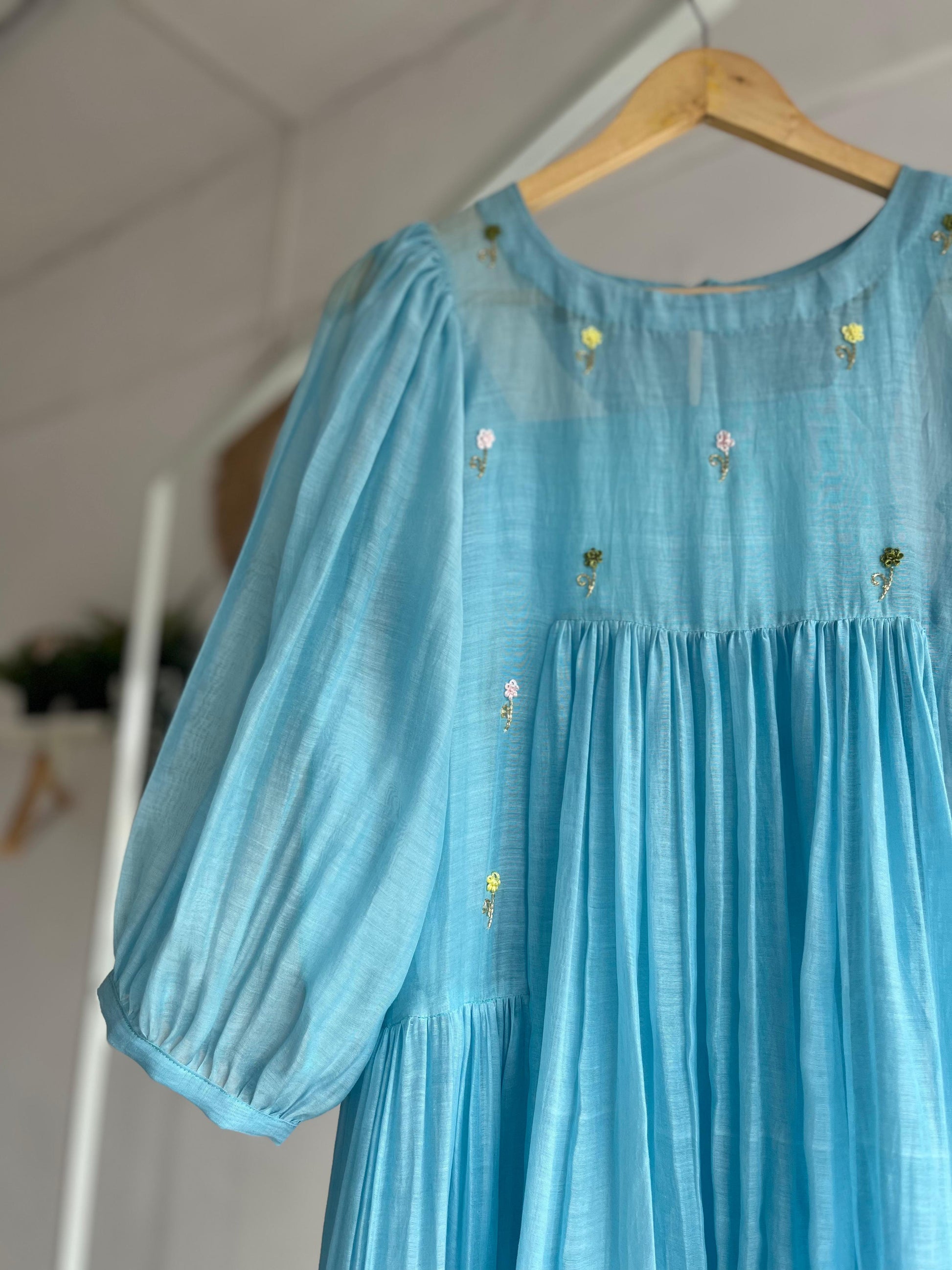 Powder blue cotton mul dress with puff sleeves, floral hand embroidery on the yoke, and a two-tiered flared hem hanging on a wooden hanger against a white background.
